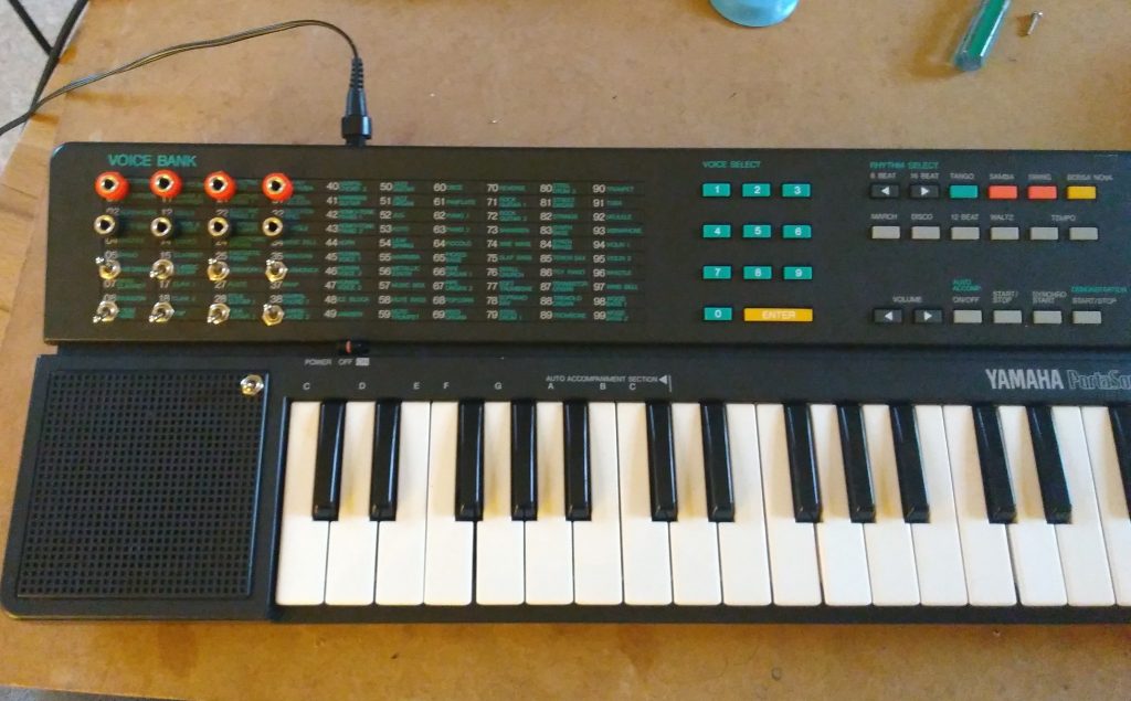 A Yamaha Portasound PSS-30 keyboard is featured, showcasing 32 black and white keys. Positioned on the left side is a built-in speaker, while above the keys lie an array of buttons and dials designated for voice selection, rhythm selection, and control settings. The label "Voice Bank" appears prominently at the top.