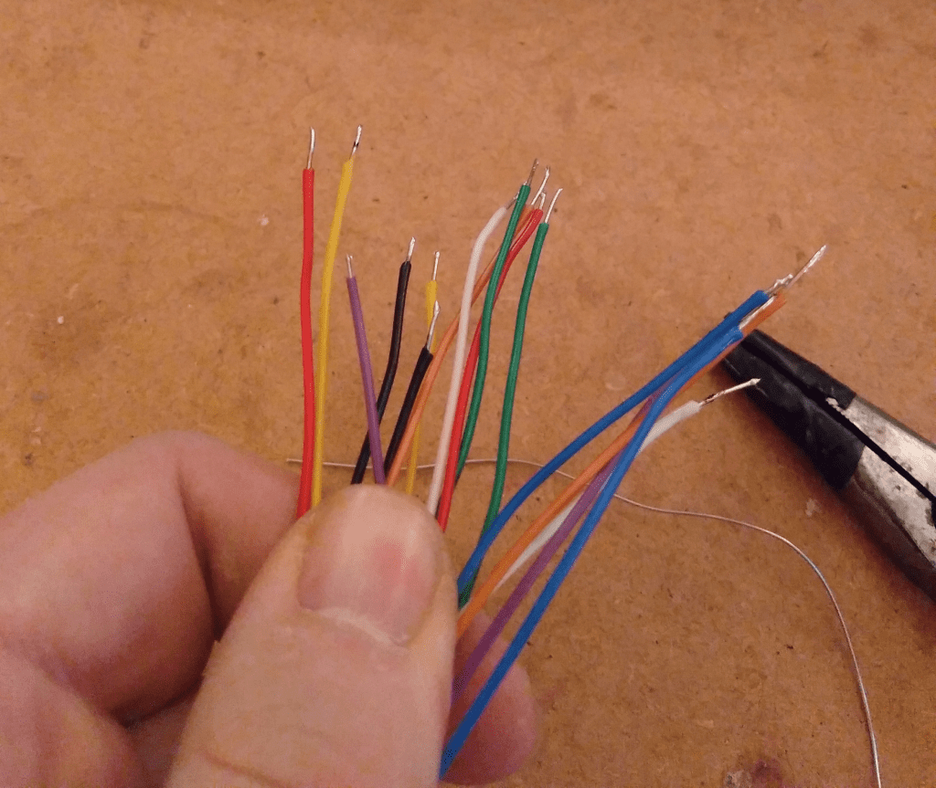 Multiple colored wires with their ends stripped are held by someone's fingers in a close-up photo. The collection includes yellow, orange, black, white, green, blue, and red wires. Nearby on a wooden surface lies a pair of needle-nose pliers.
