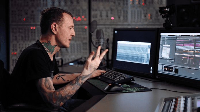Sitting at a desk in front of multiple computer monitors, Deadmau5 gestures animatedly while explaining something. The screens display audio editing software, and the background brims with various electronic equipment.