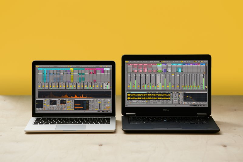 Two laptops rest side by side on a light wooden surface against a yellow background, each showcasing music production software with an array of controls, waveforms, and color-coded sections. The silver laptop on the left contrasts with the black laptop on the right, creating a visually striking setup.