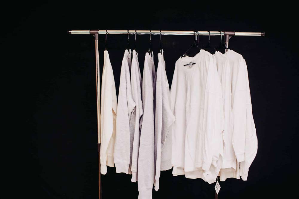 Several light-colored garments, including shirts and sweatshirts, are displayed on a clothing rack against a black background. The items, neatly hung on hangers, are arranged from left to right in varying shades of white and gray.