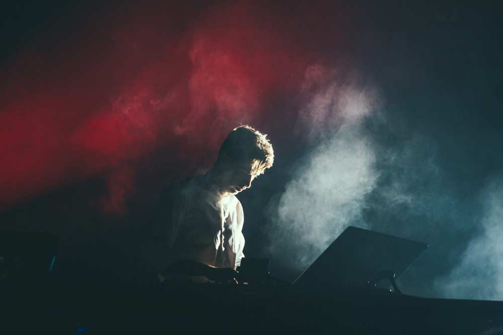 A musician stands behind a mixing console, illuminated by dramatic lighting with red and white smoke effects. The atmosphere is dark and ethereal, highlighting the intense focus of the artist.