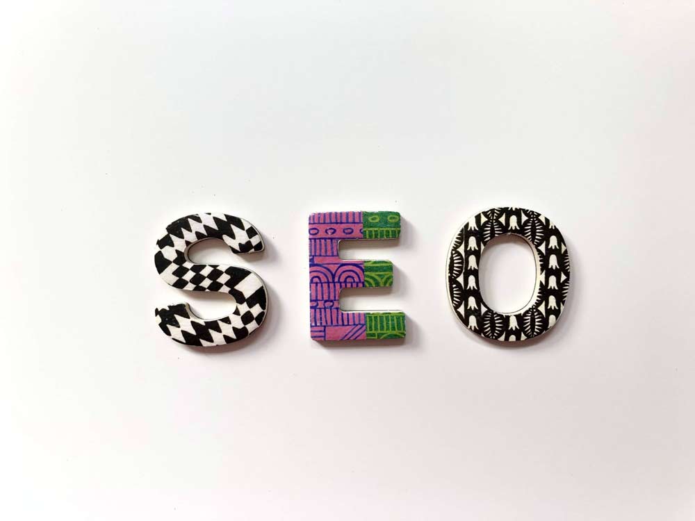 The word "SEO" is displayed using three distinct and decorated letters. The "S" presents a black and white zigzag pattern, while the "E" showcases a colorful, intricate design incorporating purple and green hues. Meanwhile, the "O" is adorned with a black and white geometric pattern.