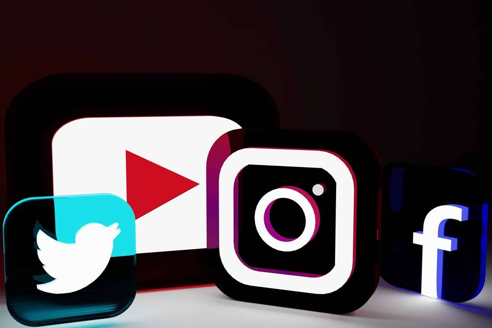 Several illuminated social media icons stand out against a dark background in this 3D composition. The design prominently features the logos of YouTube (play button), Instagram (camera), Twitter (bird), and Facebook (letter "f"), all brightly lit to capture attention. The stark contrast between the glowing icons and the dark backdrop underscores their significance in modern digital culture.