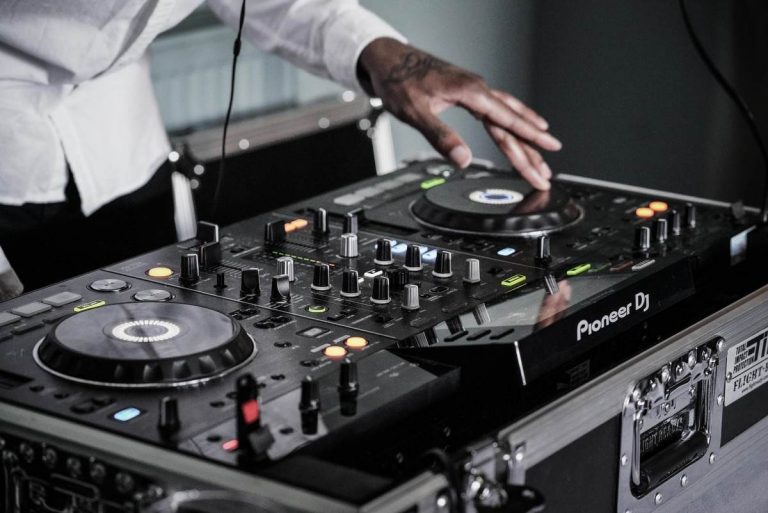 A person wearing a white shirt is mixing music using a Pioneer DJ controller. Right hand on one of the jog wheels, left hand adjusting a knob, they navigate through various buttons, knobs, and sliders. All this sophisticated equipment is housed in a sleek metal case.
