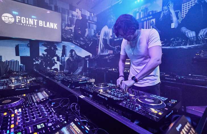 A DJ wearing a white shirt with headphones around his neck mixes music at a colorful DJ booth, surrounded by DJ equipment and mixers. Behind him are images of other DJs set against an illuminated backdrop with blue and purple lights. "Point Blank Los Angeles" is prominently displayed on a screen in the background.