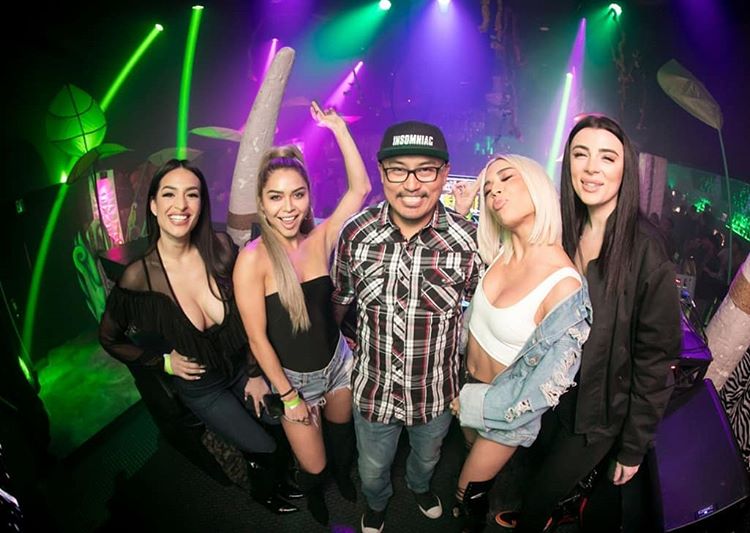 In a lively club setting with colorful lights, five people pose together. Stylish outfits are donned by the four women, who surround a man in a checked shirt and black cap. Smiles adorn everyone's faces, and one person adds an enthusiastic peace sign to the scene. The background brims with vibrant decorations, enhancing the energetic atmosphere.