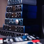 A stack of blue and black audio interface units with various knobs and connections sits in front of a blurred mixing console and speakers in the background, indicating a professional audio setup through its neat arrangement.