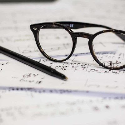 A pair of black-framed glasses and a black pen are resting on top of scattered sheets of music notation, suggesting a scene of musical composition or study. Strewn around are other signs of dedication and creativity—pages filled with intricate notes and annotations, highlighting the effort invested in crafting or understanding the music. This setting evokes a sense of quiet concentration and artistic endeavor.
