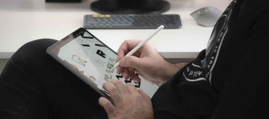 Using a stylus, a person is drawing on a tablet to create artistic letter designs. On the desk in the background, a wireless keyboard and mouse can be seen. Wearing a dark shirt, the individual seems to be seated comfortably and focused on their digital artwork.