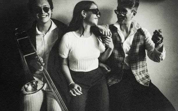 A black-and-white photo shows three people sitting closely together, all wearing sunglasses and smiling. The person on the left holds a trombone, while the one on the right sports a checkered vest. Between them, the middle person leans toward both, appearing cheerful.