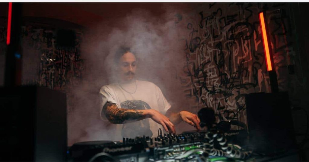 In a dimly lit room filled with fog, a DJ mixes music on turntables. Wearing a white T-shirt and showcasing tattoos on his arms, he stands against a backdrop featuring abstract graffiti art and two vertical red lights.