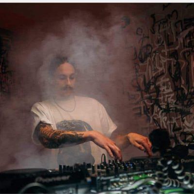 In a dimly lit room filled with fog, a DJ mixes music on turntables. Wearing a white T-shirt and showcasing tattoos on his arms, he stands against a backdrop featuring abstract graffiti art and two vertical red lights.