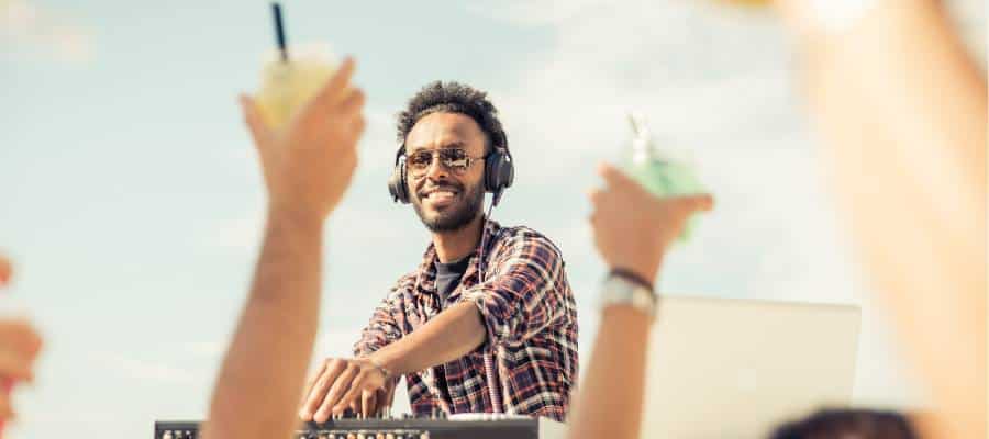 A DJ wearing headphones plays music outdoors while smiling. He is dressed in a plaid shirt and sunglasses. In the foreground, two people raise drinks, creating a lively atmosphere under a clear sky.
