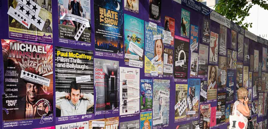 A wall covered in colorful posters and advertisements for various performances and events, including comedy shows and musical acts, forms the backdrop. A person stands on the right side, looking at the posters. In the top right corner, a green tree partially shades the scene.