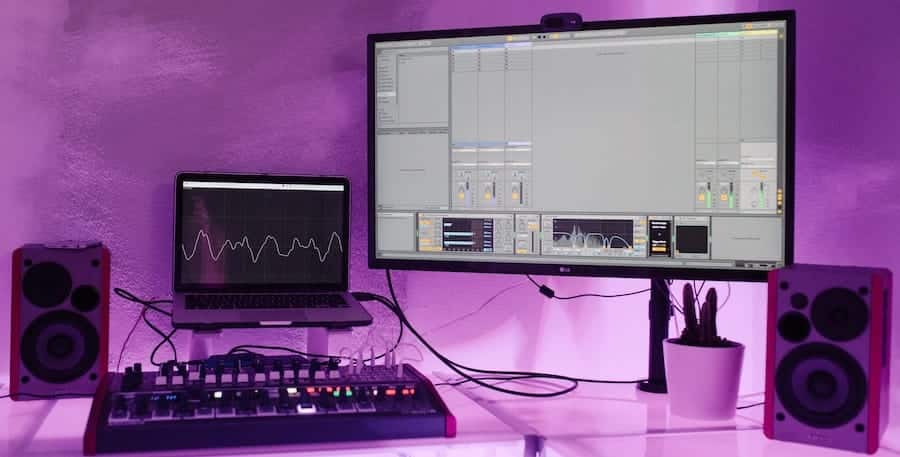 A music production setup with a desktop monitor displaying Ableton, a laptop showing sound waves, two speakers, a MIDI controller, and a small potted plant, all on a desk with a purple lighting backdrop.