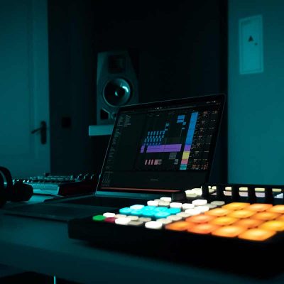 A laptop on a desk in a dimly lit room displays Ableton, accompanied by headphones, an audio mixer, and a MIDI controller with brightly lit pads. In the background, a large speaker adds to the ambiance.