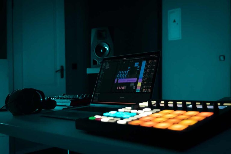 A laptop on a desk in a dimly lit room displays Ableton, accompanied by headphones, an audio mixer, and a MIDI controller with brightly lit pads. In the background, a large speaker adds to the ambiance.