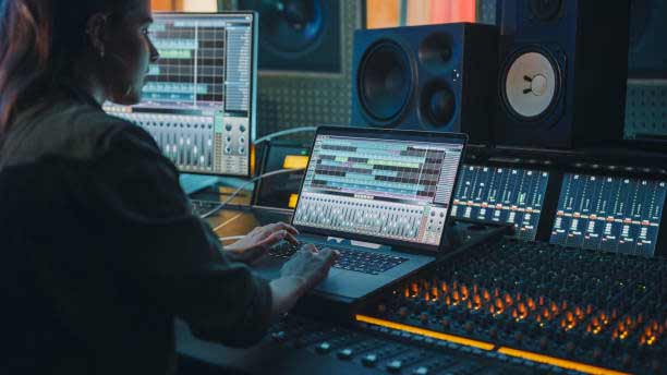 A person works on audio editing software on a laptop in a dimly lit music studio. Surrounding them are large speakers, various mixers, and sound control panels with numerous knobs and buttons, creating an environment rich with professional audio equipment.