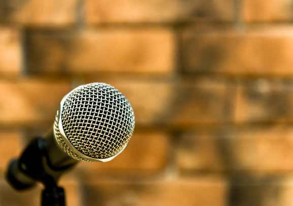A silver mesh grille on a microphone is featured in a close-up shot, set against an orange brick wall and mounted on a black stand.
