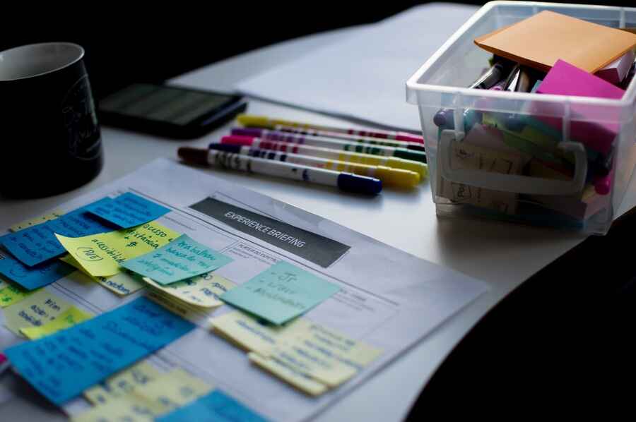 A white desk holds a sheet of paper adorned with colorful sticky notes and captions labeled "experience briefing." Nearby, assorted markers, a plastic container filled with more sticky notes, and a mug contribute to the creative workspace. A portion of a notebook peeks from the background, adding to the organized chaos.