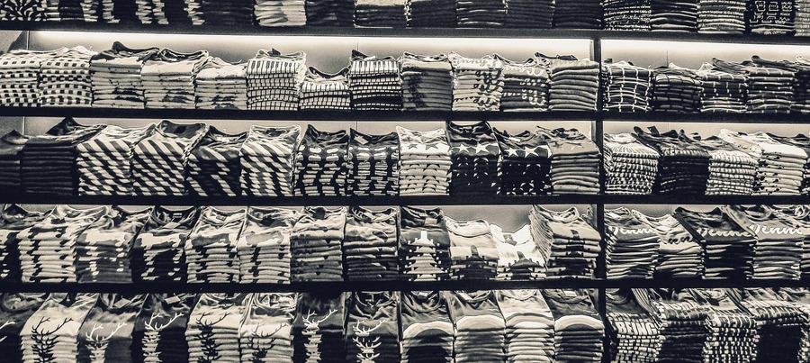 A neatly organized display of folded graphic T-shirts on shelves in a store includes various patterns and designs, primarily featuring stars, stripes, and other geometric shapes. Captured in black and white, it exudes a modern and sleek feel.