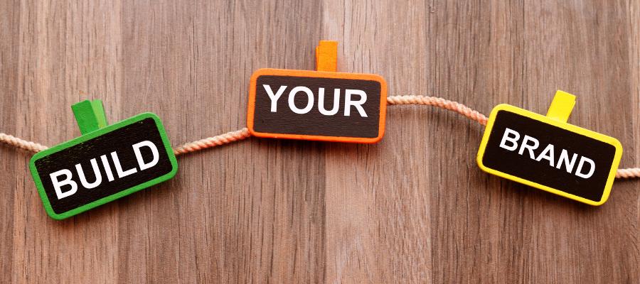 Three small rectangular signs hang on a rope against a wooden background. The signs are labeled "BUILD," "YOUR," and "BRAND," from left to right. Each sign is in a different color: green, orange, and yellow, respectively.