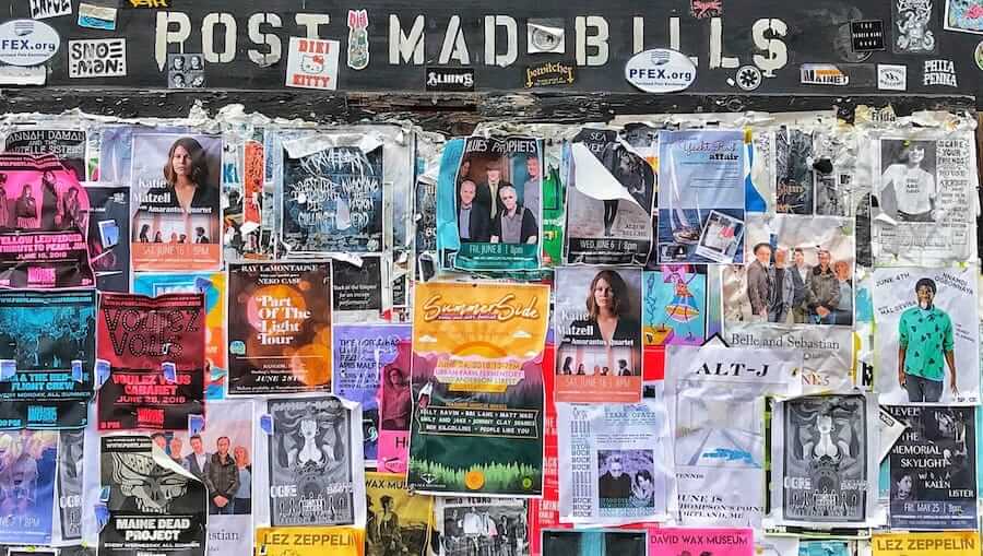 A crowded bulletin board is covered with a variety of colorful posters and flyers advertising concerts, events, and local performances. Some posters are partially torn or overlapped.