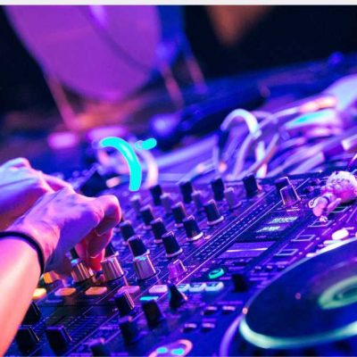 Bathed in vibrant blue and purple lights, a close-up captures a DJ's hands deftly adjusting knobs on a mixing console. Various pieces of audio equipment, such as turntables and cables, contribute to the energetic atmosphere of the scene.